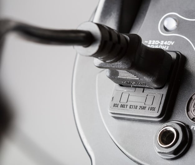A detailed close-up of an electric vehicle's charging port, showing the connector plugged in and the vehicle's brand logo prominently displayed. The background includes a blurred view of a residential garage.